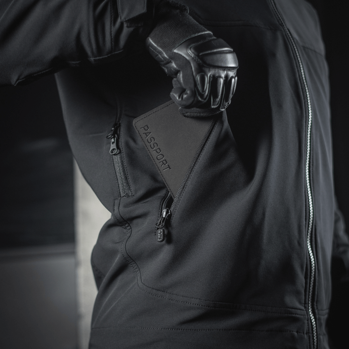 Person i sort jakke, der holder M‑Tac Pascover med "Passport"-tryk, praktisk til rejser.