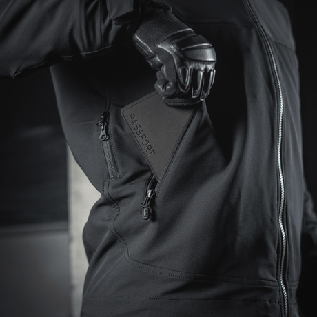 Person i sort jakke, der holder M‑Tac Pascover med "Passport"-tryk, praktisk til rejser.