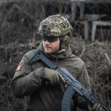 Person iført M‑Tac Vilha Hjelmcover FAST i Multicam, holder våben i ujævnt terræn.