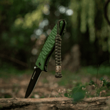 Den M-Tac Viper Lanyard Scandinavian i flettet paracord med Viper perle, ved en lommekniv i naturen. Stilfuldt stilfuldt tilbehør.