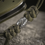 M-Tac Zeus Stainless Steel Lanyard med flettet paracord og dekorativ metalperle på en kniv, perfekt til hurtig adgang.