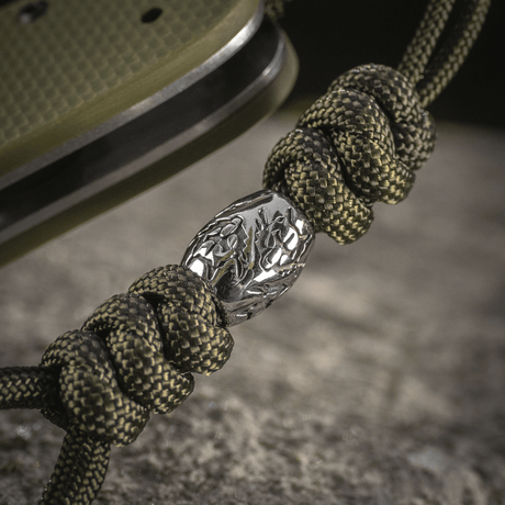 M-Tac Zeus Stainless Steel Lanyard med flettet paracord og dekorativ metalperle på en kniv, perfekt til hurtig adgang.