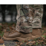 M-Tac Feltbukser NYCO i camouflagemønster, fremstillet af slidstærkt NYCO-stof, vises med kraftige støvler på skovbund.