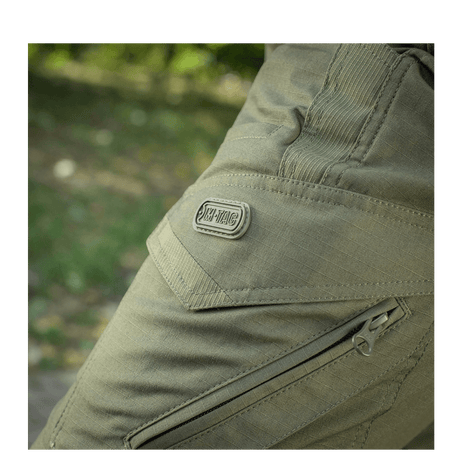 Detalje af M-Tac Bukser Aggressor Dame Flex i grønt, viser robust lynlås og logo, ideel til træning og udendørs aktiviteter.