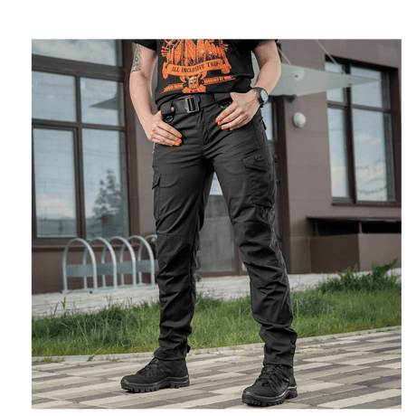 Model iført M-Tac Bukser Aggressor Dame Flex foran en bygning, sort og taktisk design, velegnet til forskellige udendørs aktiviteter.