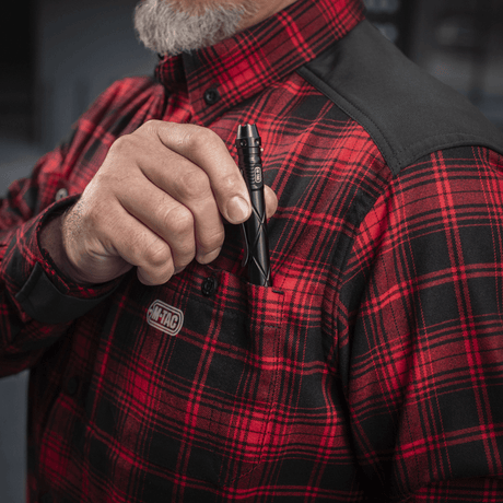 Mand iført M-Tac Redneck Skjorte i rød plaid og sort forstærkning, holder en lommelygte op af brystlommen.