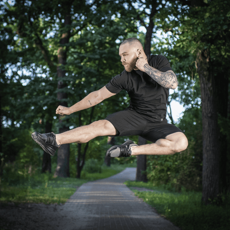 Mand i M-Tac Shorts Casual Fit Cotton udfører et spring i parken, iført afslappet sort tøj, fremstillet af åndbart 100% bomuld.