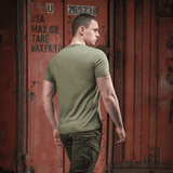 Person iført armygrøn M-Tac T-shirt Viking med ryggen til, gående forbi container i bymiljø.