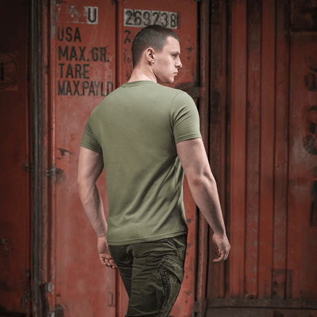 Person iført armygrøn M-Tac T-shirt Viking med ryggen til, gående forbi container i bymiljø.