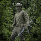 Person iført M-Tac Division Gen.II Windblock Fleecejakke, designet til maksimal varme og vindbeskyttelse, vandrer i skovområde.