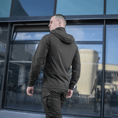 Mand iført sort M-Tac Flash Jakke, perfekt til vandreture og hverdagsbrug, set bagfra foran moderne bygning.