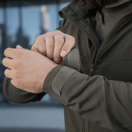 Person justerer ærmen på en grøn M-Tac Flash Jakke i et udendørs miljø.