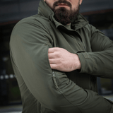 Mand bærer grøn M-Tac Flash Jakke, letvægts og alsidig til udendørs aktiviteter, ses justere jakken uden velcrofelter.