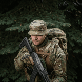 Camouflagenet mand med M-Tac Panama Hat Gen.II NYCO, taktisk uniform og rygsæk i skovmiljø.