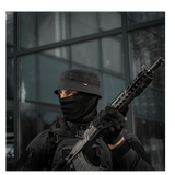 Person wearing tactical gear with a hat and face covering, holding a rifle, standing outside a building.