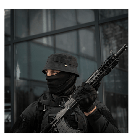 Person wearing tactical gear with a hat and face covering, holding a rifle, standing outside a building.