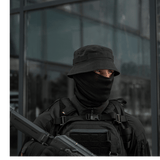 Mand iført taktisk tøj med sort hat, maske og rygsæk foran glasbygning.