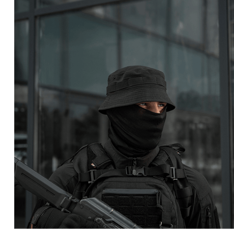 Mand iført taktisk tøj med sort hat, maske og rygsæk foran glasbygning.