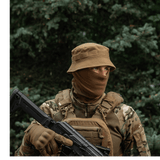 Person i camouflage med hat og maske i skov, bevæbnet med gevær.