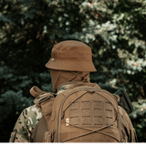 Person i taktisk camouflagedragt og rygsæk iført M-Tac Panamahat Gen. II Flex Rip-Stop i skovmiljø.