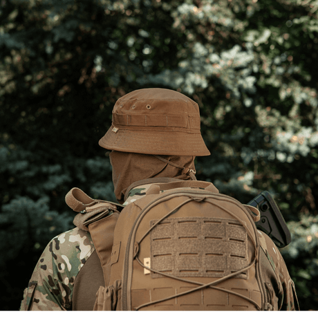 Person i taktisk camouflagedragt og rygsæk iført M-Tac Panamahat Gen. II Flex Rip-Stop i skovmiljø.