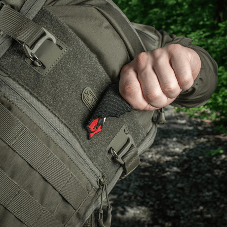 Person trækker en M-Tac Patch Husaria 3D PVC morale patch ud af en taktisk rygsæk, viser vingede husar design mod grøn skov baggrund.