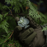 M-Tac Ace of Spades 3D PVC Patch holdt af en taktisk handskeklædt hånd i skovmiljø, ideel til militære og outdoor-aktiviteter.