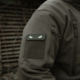M-Tac Cat Eyes 3D PVC Patch på jakkeærme i taktisk miljø, viser lysreflekterende og fluorescerende design, fastgjort med Velcro.