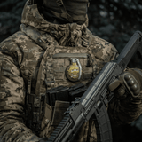 Soldat med camoufleret jakke bærer M-Tac Patch M67 Granat 3D PVC på vesten, humoristisk emoji-design, taktisk militærudstyr.