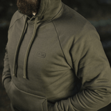 Mand iført M-Tac Cotton Raglan Hard Hoodie i oliven, med raglanærmer, hætte og kængurulomme. 100% bomuld og fleeceforet til varme.