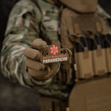 M-Tac Patch Viking Evac i soldatens hånd, viser Odin will wait paramedicine motiv. Camouflage jakke og taktisk vest baggrund.