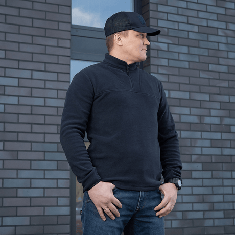 M-Tac Delta Polartec Fleecejakke i sort, fremstillet af Polartec®-fleece, bæres af en mand foran en murstensvæg.