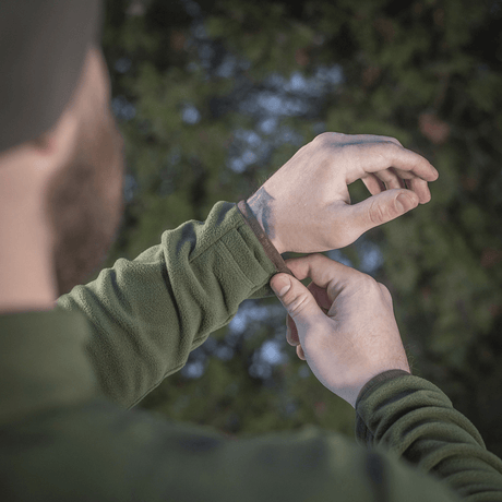 Mand justerer manchetten på en M-Tac Delta Polartec Fleecejakke i en skov, som viser jakkens funktionelle design og materiale.