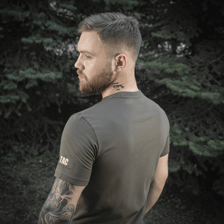 Mand bærer M-Tac Sniper T-shirt i skovmiljø, viser holdbar og komfortabel pasform af 93% bomuld og 7% elastan, ideel til udendørs brug.