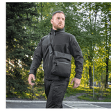 Mand iført M-Tac Konvert Taske Elite, udendørs i park, iført sort tøj og gående i naturligt lys, taskens komfort og stil vises.