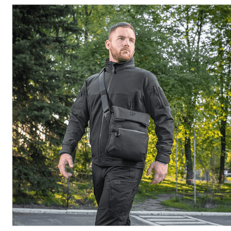 Mand iført M-Tac Konvert Taske Elite, udendørs i park, iført sort tøj og gående i naturligt lys, taskens komfort og stil vises.