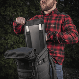 En person åbner M-Tac Rygsæk Gen. II Elite – Small med indsat rygstivhed, iført rødternet skjorte, udendørs.