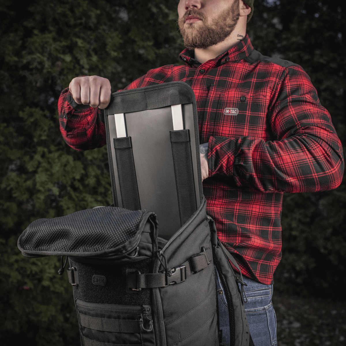 En person åbner M-Tac Rygsæk Gen. II Elite – Small med indsat rygstivhed, iført rødternet skjorte, udendørs.