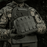 M-Tac organizer pouch Elite holdt af en taktisk klædt person, fastgjort til vest med M.O.L.L.E.-system, velegnet til førstehjælp og EDC-udstyr.