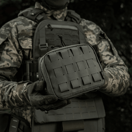 M-Tac organizer pouch Elite holdt af en taktisk klædt person, fastgjort til vest med M.O.L.L.E.-system, velegnet til førstehjælp og EDC-udstyr.