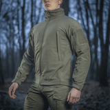 Mand iført M-Tac Soft Shell Jakke i skovmiljø, designet til at beskytte mod dårligt vejr med indre polyuretan-membran og fleece-lag.