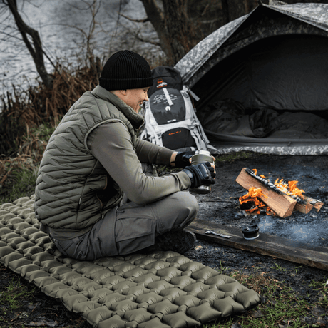 Mand sidder på M-Tac Oppustelig Liggeunderlag 195x60 tæt ved lejr, komfortabel camping ved bål