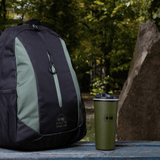 M-Tac Termokop 450 ml med Ventil står ved siden af en rygsæk på et bord i en skovscene.