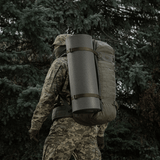 Person med camouflage, der bærer rygsæk med M-Tac Liggeunderlag 20 mm (60x180 cm) i skovmiljø.