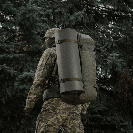 Person med camouflage, der bærer rygsæk med M-Tac Liggeunderlag 20 mm (60x180 cm) i skovmiljø.