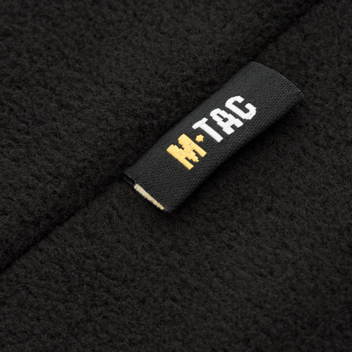Nærbillede af M-Tac Delta Fleece Jakke etikette, sort og gul logo, fleece materiale, ideel til udendørs og kolde vejrforhold
