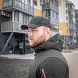 Mand iført en sort M-Tac Mesh Flex Ripstop Baseball Cap foran bybygninger, viser udendørs holdbarhed og stil.