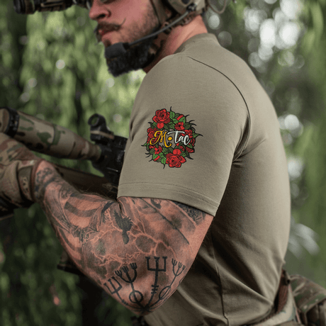 Mand med tatovet arm iført M-Tac T-Shirt Motanka, holder en riffle i naturlig omgivelser, fremhæver slavic inspireret design.