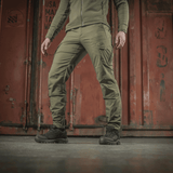 M-Tac Taktiske Bukser Aggressor Gen. II Flex i grøn farve mod en industriel baggrund