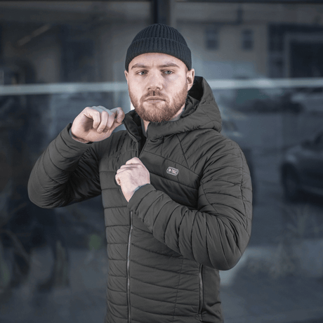 Mand iklædt M-Tac Jarl Jakke foran bygning, opkvikker jakken og ser betragtende fremad i urban omgivelse.